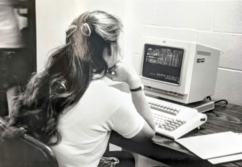 Black and white photo of woman sitting at an old desktop computer